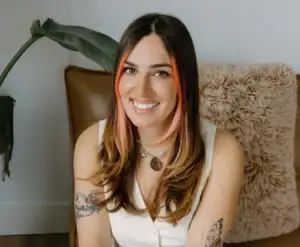 Micah Litowitz wearing a white top and jeans, sitting on a couch and smiling at the camera