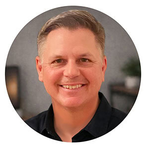 A professional headshot of Corey Gasman, a web designer, smiling and wearing a grey polo shirt against a neutral, light-colored background.