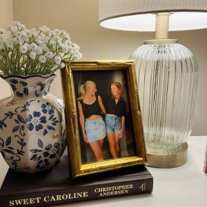 Gold‑toned nightstand styled with the book ‘Sweet Caroline,’ a blue and white vase, gold photo frame of two friends, and a lamp — chic and personal bedside table decor ideas from By Paige Nicole.