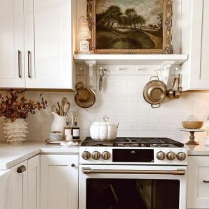 White kitchen and stove decorated with brass pots, pans, and a pumpkin kettle — cozy fall‑inspired top of stove decor ideas from By Paige Nicole.