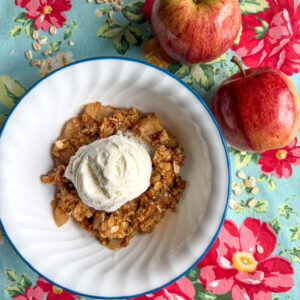 Bowl of fresh milled flour apple crisp on a bright colored kitchen placemat with two apples on the side.