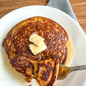 Pumpkin pancakes with fresh milled flour with a bite taken out on a fork. Pancakes are on a white plate on a wooden table with a blue and white striped cloth napkin next to them.