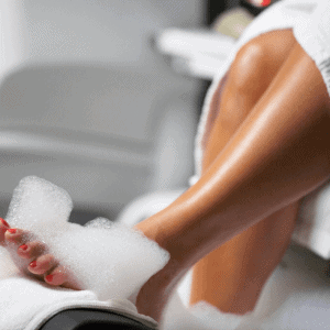 A close-up of a woman's legs and feet during a luxurious pedicure session. She is sitting with her feet soaking in a bubbly foot bath, with her toenails painted a vibrant red. She is wrapped in a white robe, creating a relaxing and pampering atmosphere. The background is softly blurred, focusing attention on the pedicure.