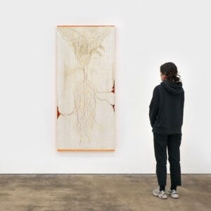 A person in dark clothing stands in a gallery at the Xin-Liu exhibition at Frieze London 2025, viewing a large vertical artwork on a white wall. Abstract, pale lines and shapes resembling roots or branches stretch across a light background.