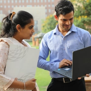 Two people outdoors, one holding a folder, the other using a laptop, with a building in the background.