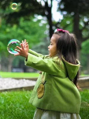 Young child wearing a green coat, reaching out to catch a bubble outdoors.