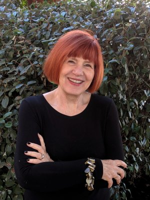 A portrait of a woman with vibrant red hair smiling warmly, dressed in a black top. She is standing outdoors with a backdrop of dense, green foliage. Her arms are crossed, and she wears a distinctive bracelet with key charms.