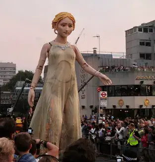 A photograph of an over-sized human puppet of light skinned, blonde puppet in a white gown, parading during the Cultural Olympiad Carnval in the London 2012 Games.