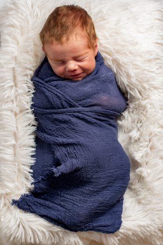 Newborn baby swaddled in a dark blue blanket, lying on a white, fluffy rug.