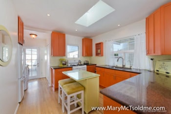 Bright kitchen with orange cabinetry, a skylight, dark countertops, and a small yellow island with seating.