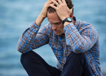 A man in a plaid shirt sits by the water looking distressed, symbolizing stress., tags: gut–brain habits mental - pexel