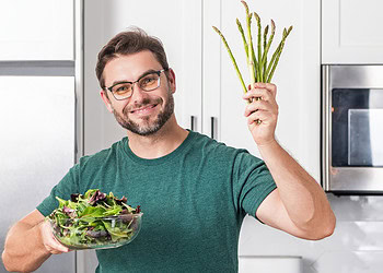 Fresh green and purple salad vegetables and arugula in a glass bowl, man holding a bunch of asparagus and smiling in a modern kitchen, healthy eating, plant-based diet, nutritious lifestyle, PsyPost news.