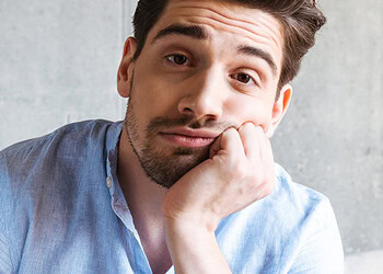 Thoughtful young man with a friendly expression, sitting indoors with a concrete wall background, illustrating themes of psychology, mental health, and emotional well-being.
