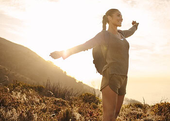 A woman enjoying outdoor nature hike, spreading arms joyfully against a scenic coastal background at sunrise.