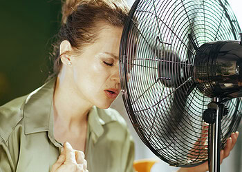 Sweat-drenched woman cooling off with electric fan outdoors on hot day, showcasing mental health, heat stress, and environmental factors affecting well-being, in a natural outdoor setting.