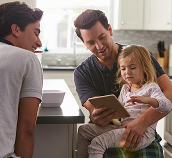 A man in pyjamas with a young girl also in pyjamas sitting on his knee whilst they are both looking at a tablet he is holding and another man is sitting with them.