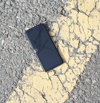 A close-up view of a cracked smartphone lying on a sunlit asphalt road.