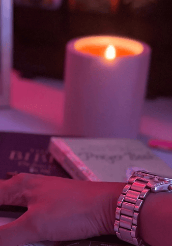 A serene devotional setup featuring an open Bible highlighted in pink lighting, with a hand resting gently on its pages. A lit candle, a "Prayer Journal," a mug with the letter "T," and a framed photograph in the background create a peaceful atmosphere. The image symbolizes faith and trust in manifesting what’s already destined for you.