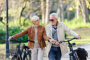 An older couple walks their bicycles down a tree-lined path in a sunny Raleigh park