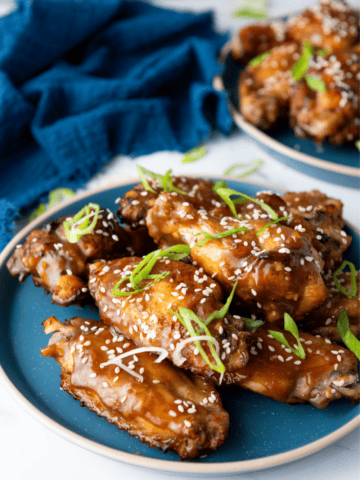 A close-up view of sticky Chinese Chicken Wings garnished with sesame seeds and green onions on a blue plate, set against a dark blue cloth background.