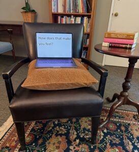 Photo of a laptop on a chair in a therapy office. Text on the laptop screen says, "How does that make you feel?"