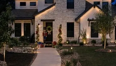 Holiday-decorated Central Texas home with warm landscape lighting, illuminated evergreens, and a festive wreath at the front entrance.