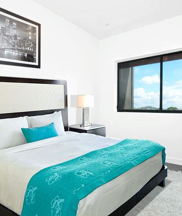 Ein helles, modernes Schlafzimmer mit einem großen Bett, weißen Laken und einem türkisfarbenen Überwurf. Links hängt ein gerahmtes Schwarz-Weiß-Bild einer Skyline, und rechts steht ein Nachttisch mit einer Lampe neben einem Fenster mit Blick auf den blauen Himmel.