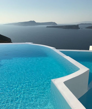 Ein atemberaubender Infinity-Pool mit strahlend blauem Wasser erstreckt sich vor einer weiten Meereslandschaft mit mehreren Inseln und einem klaren Himmel. Die Architektur des Pools ist modern und weiß.