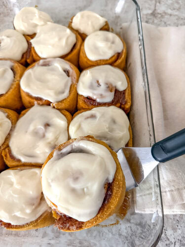 Pumpkin Cinnamon Rolls in a baking dish on top of a kitchen counter with a kitchen spatula lifting one cinnamon roll out of the pan.