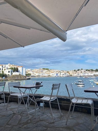 Idyllic seaside café terrace with views of calm waters and white buildings under a partly cloudy sky, ideal for relaxing dining experiences.