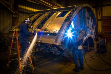 Welding operator welding on large piece of vibrating equipment with another person grinding