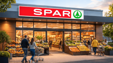 **Alt text:** A realistic exterior view of a modern SPAR supermarket at sunset, showing the red SPAR sign with a green tree logo above large glass windows. Shoppers are entering and exiting the store while fresh fruits and vegetables are displayed outside in crates, creating a busy