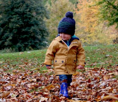 Kind mit Jacke und Mütze steht auf einer Wiese auf der viel Laub liegt