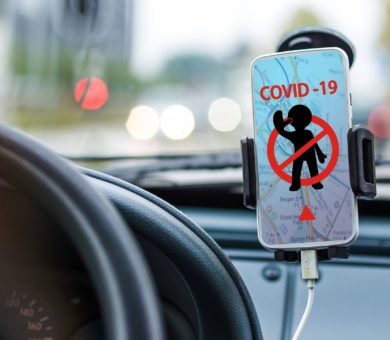 Ein Smartphone, das in einer Halterung neben einem Autolenkrad befestigt ist zeigt ein durchgestrichenes Männchen. Darüber steht COVID-19.