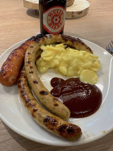 Lecker gegrilltes: 2 SDK Würste und eine rote Wurst mit Kartoffelsalat und einem Wulle im Hintergrund