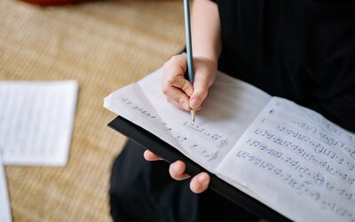 Person's hands write musical notes with a pencil on opened sheet music in a binder