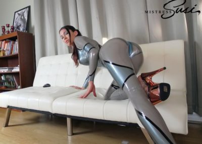 Woman in silver latex bodysuit posing on a white sofa in a modern room.