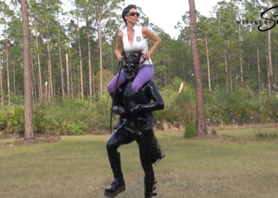 Woman in fetish outfit standing on a man in an outdoor forest setting.