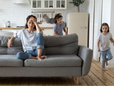 Sonja hat ein Mama-Burnout, Mutter sitzt auf Sofa, zwei Kinder springen rundherum.