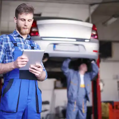Auto mechanic reviewing service data on a tablet while another mechanic works under a raised vehicle