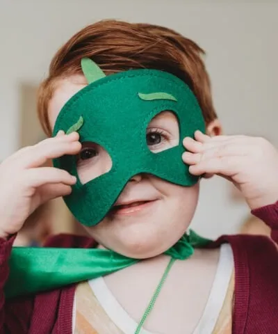 Young child dressed as a superhero with a green mask and cape, adjusting their costume.