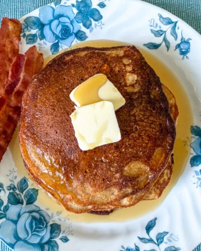Landscape view of a stack of einkorn flour pancakes on a blue floral plate, served with butter, maple syrup, and two strips of bacon