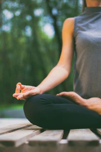 A person sitting cross-legged outdoors on a wooden surface, practicing meditation or yoga. Their hand rests on their knee in a relaxed position, with fingers forming a mudra, set against a blurred background of greenery.