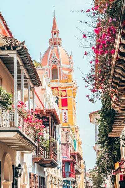 Flower-covered balconies in the foreground lining a street, with the yellow and pink tower of Cartagena, Colombia's cathedral rising above.
