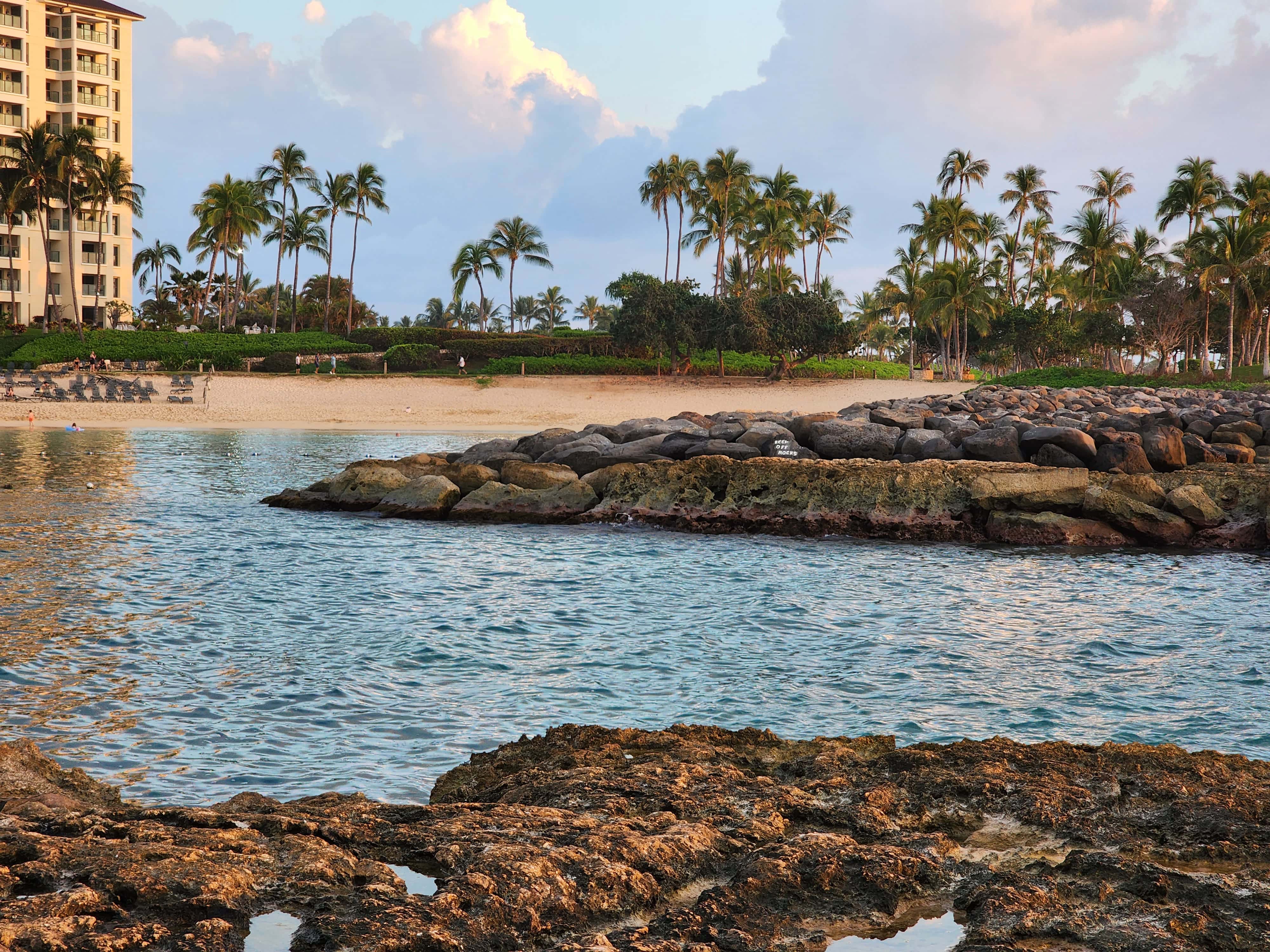 A serene beach in Kapolei, Oahu, Hawaii, featuring calm turquoise waters, rocky outcroppings, and a backdrop of palm trees and a coastal building under a pastel sky at sunset. The image is used as a featured image for the blog post "What is Sensory Overload?"