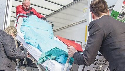 Menschen heben ein Krankenhausbett, das voll Bezogen und mit Bettwäsche ausgestattet ist in einen Transporter.