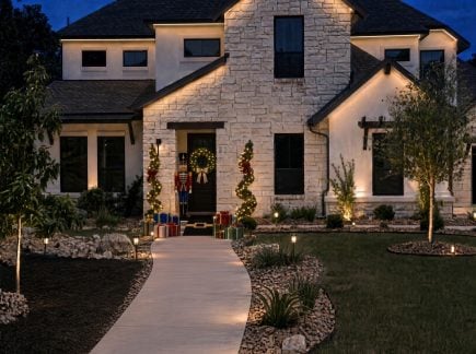 Holiday-decorated Central Texas home with warm landscape lighting, illuminated evergreens, and a festive wreath at the front entrance.