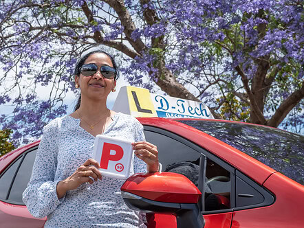 Happy student standing with P-plate under violet blooming trees next to red training car