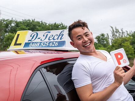 Male student leaning back on red car smiling holding fresh P-plate in celebration
