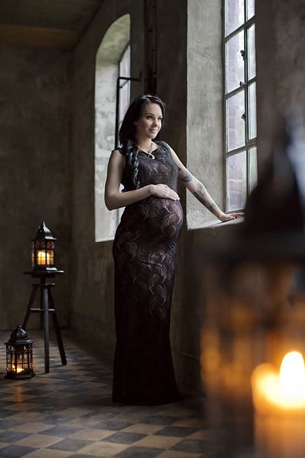 Elegant gravidfotografi av gravid kvinne i svart kjole som hviler hånden på magen, inne i et rom med stort vindu, skaper en varm og vakker gravidfotoseanse.
